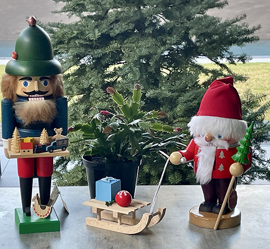 Ein deutscher Nussknacker, ein hölzerner Schlitten mit einem blauen Geschenk und einer roten Schneeflocken-Weihnachtskugel, gezogen von einem Weihnachtsmann-Nussknacker, steht auf einem Tisch vor einem Tannenbaum.