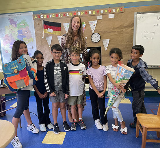 7 junge Schüler, die in einer Reihe in ihrem Klassenzimmer stehen, das einen blauen Boden hat. Sie lächeln alle. Eine Lehrerin steht in der Mitte hinter ihnen. Sie stehen alle vor einer Wand mit einer Deutschlandkarte, einer Flagge, einer kleinen Uhr und einer Tafel.
