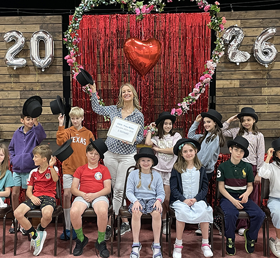 Klassenfoto der Klasse von 2026 von 13 jungen Schülern, die in Stühlen sitzen und schwarze Zylinder tragen, um das Schuljahr zu feiern. Die Lehrerin steht in der Mitte dahinter und hält ihren Zylinder in der rechten Hand erhoben über ihren Kopf. Im Hintergrund schweben silberne Ballons in Form der Jahreszahlen 2, 0, 2, 6 vor einer hölzernen Lamellenwand.